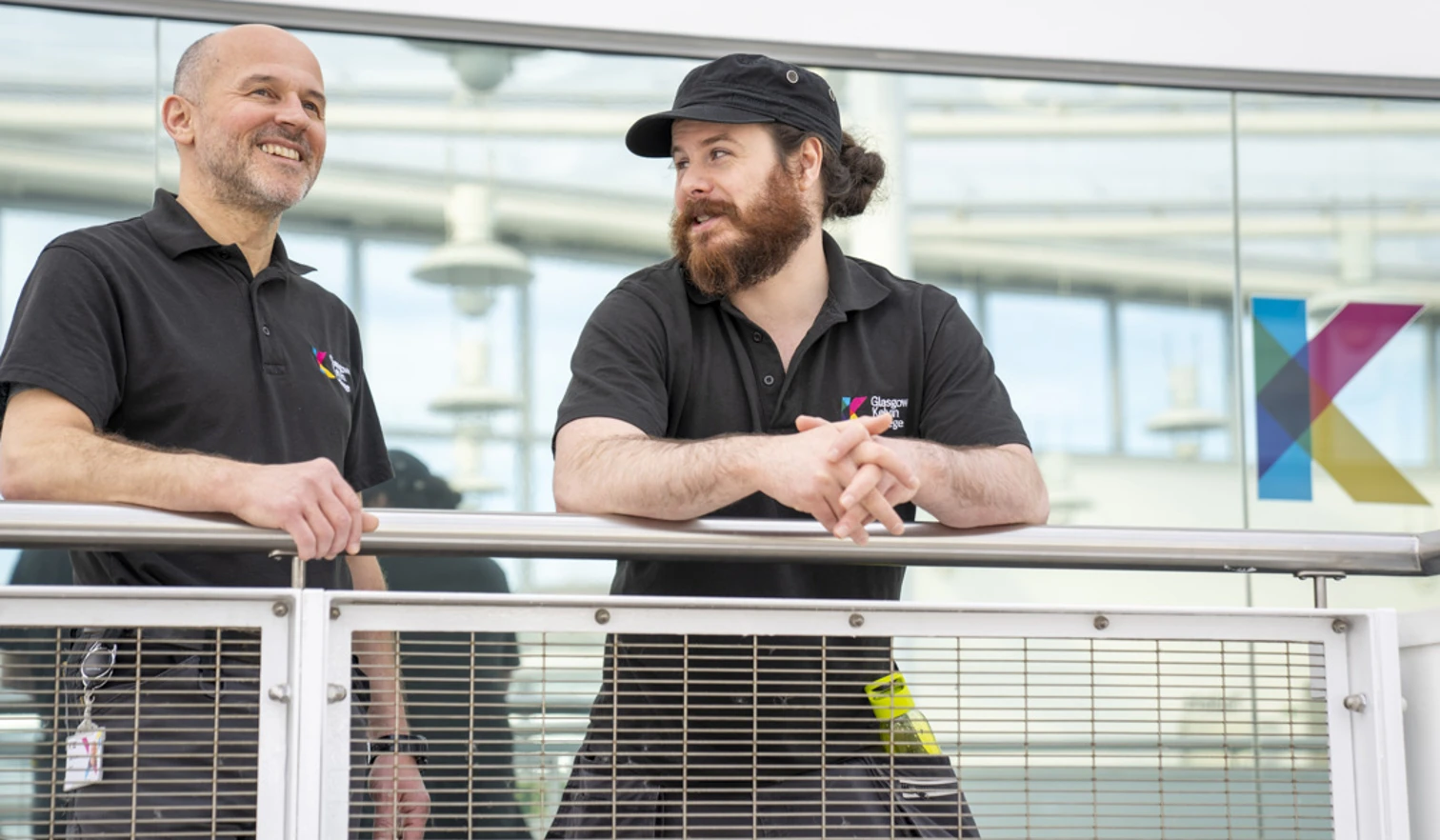 Two Glasgow Kelvin College estates staff standing by a balcony railing, smiling and talking in uniform near a window with the college logo Two Glasgow Kelvin College estates staff standing by a balcony railing, smiling and talking in uniform near a window with the college logo
